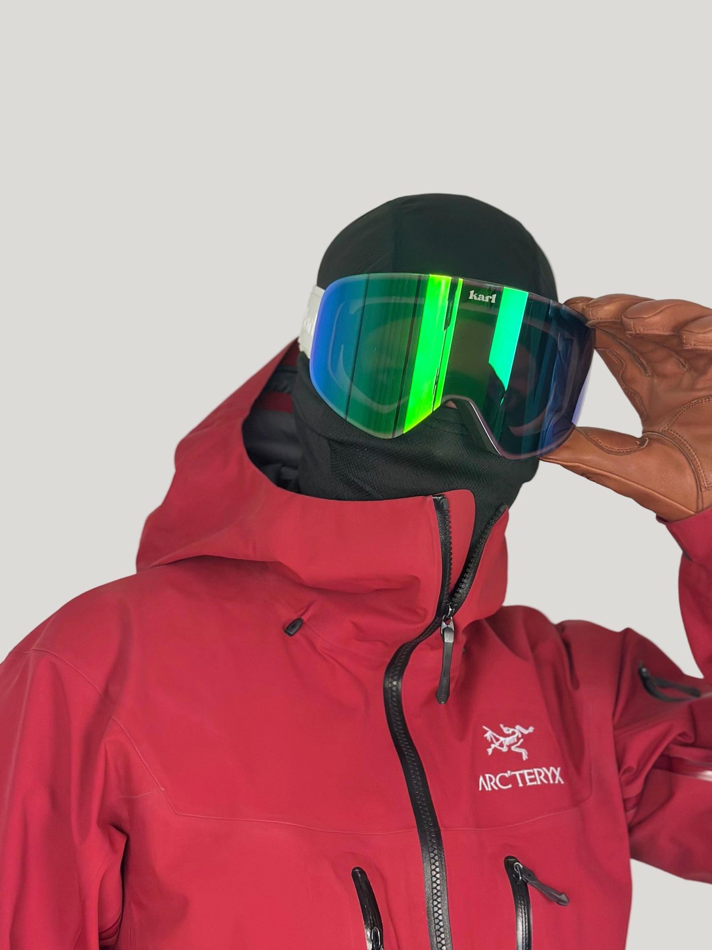 Person wearing a red jacket with a hood, holding green goggles above their head against a plain background